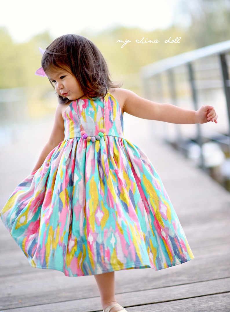 May include: A young girl wearing a colorful, abstract print dress with a beaded necklace. The dress has a gathered skirt and a sleeveless bodice. The girl is standing on a wooden walkway with the text "my china doll" in the background.