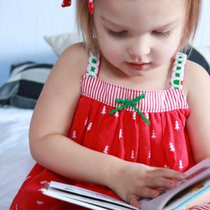 Pu&ograve; includere: Una bambina con un vestito a righe rosse e bianche con un fiocco verde e alberi di Natale bianchi stampati su di esso, sta leggendo un libro. &Egrave; seduta su un letto bianco con un cuscino a righe bianche e nere dietro di lei.