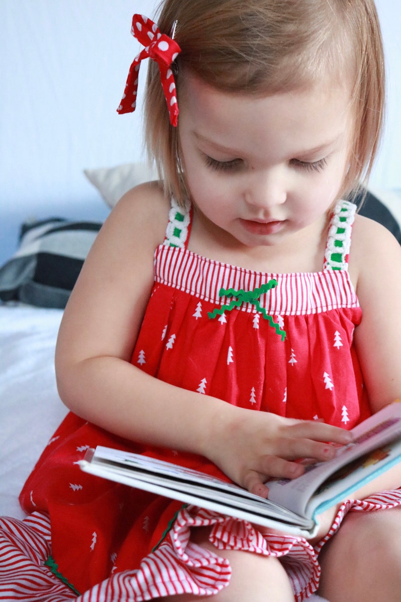 Pu&ograve; includere: Una bambina con un vestito a righe rosse e bianche con un fiocco verde e alberi di Natale bianchi stampati su di esso, sta leggendo un libro. &Egrave; seduta su un letto bianco con un cuscino a righe bianche e nere dietro di lei.