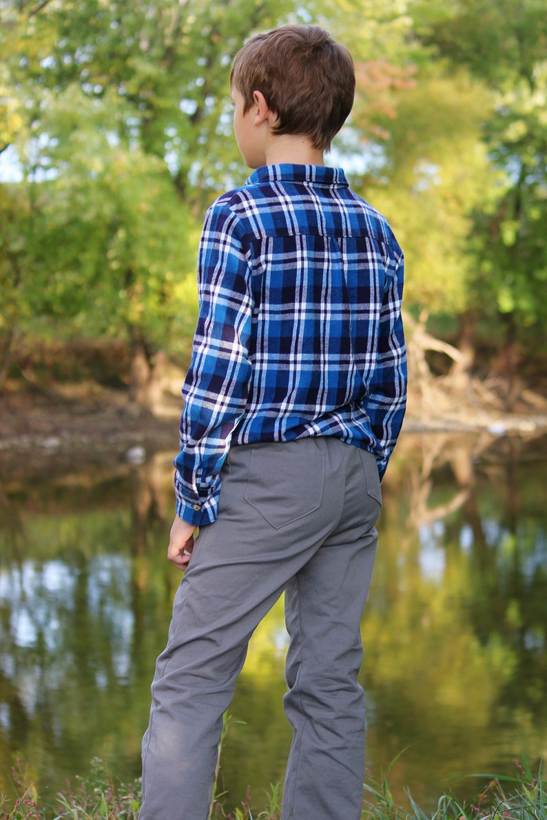 Op de afbeelding: Een jong persoon draagt een blauw-wit geruit overhemd en een grijze broek en staat aan de rand van een waterpartij. De persoon staat met de rug naar de camera en kijkt naar het water.
