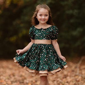 May include: A young girl wearing a dark green dress with a floral pattern. The dress has a tan-colored trim and a matching belt. She is wearing brown shoes and white socks. The background is a blurred view of a forest.