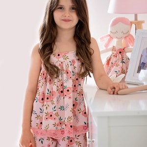 Pu&ograve; includere: Una bambina che indossa un pigiama rosa con stampa floreale e bordino in pizzo. Il set comprende una canotta e pantaloncini. La bambina &egrave; in piedi accanto a una cassettiera bianca con una lampada rosa e una bambola.