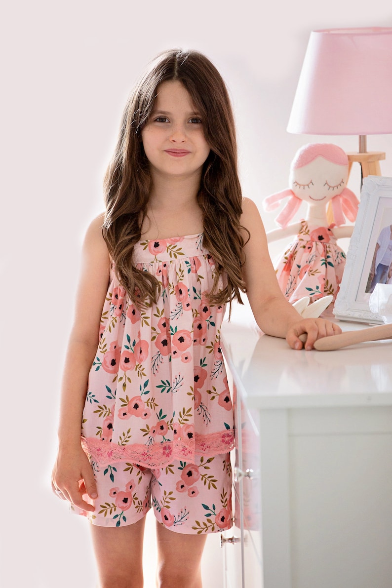 Pu&ograve; includere: Una bambina che indossa un pigiama rosa con stampa floreale e bordino in pizzo. Il set comprende una canotta e pantaloncini. La bambina &egrave; in piedi accanto a una cassettiera bianca con una lampada rosa e una bambola.