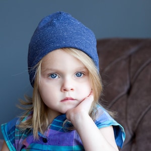 May include: A young person with long blonde hair wearing a blue beanie and a plaid shirt. They are looking at the camera with a thoughtful expression.
