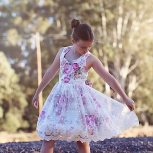 May include: A young girl wearing a white dress with pink floral print and lace trim. The dress has a sweetheart neckline and a full skirt. The girl is standing in a grassy field with trees in the background.