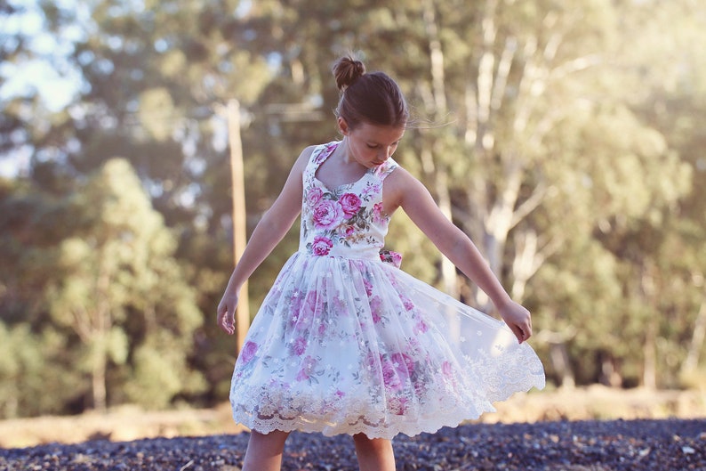 May include: A young girl wearing a white dress with pink floral print and lace trim. The dress has a sweetheart neckline and a full skirt. The girl is standing in a grassy field with trees in the background.
