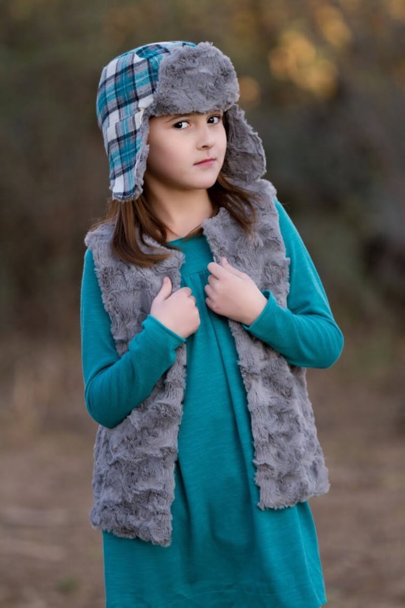 May include: A young person wearing a teal blue long-sleeved shirt with a gray faux fur vest. They are wearing a plaid and gray trapper hat with ear flaps.