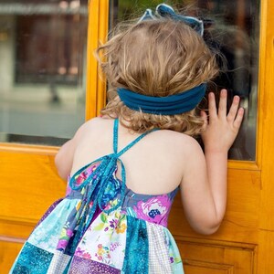 May include: A young girl wearing a colorful patchwork dress with a teal blue headband, looking out a window.