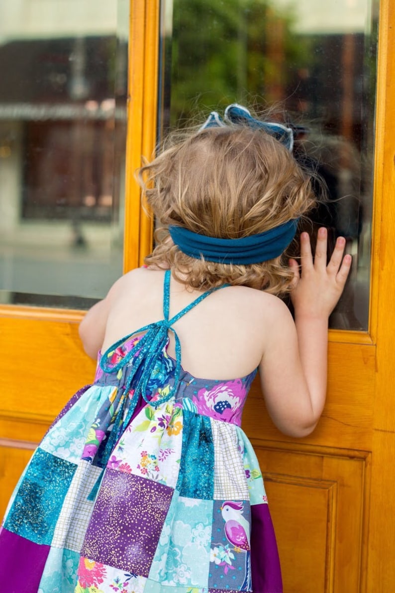 May include: A young girl wearing a colorful patchwork dress with a teal blue headband, looking out a window.