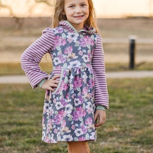 May include: A young girl wearing a purple and white striped long-sleeved hooded dress with a floral print. The dress has a green trim and pockets. She is wearing brown cowboy boots.