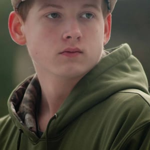 May include: A young person wearing a green hooded sweatshirt with a camouflage pattern on the inside of the hood. They are also wearing a brown newsboy cap.