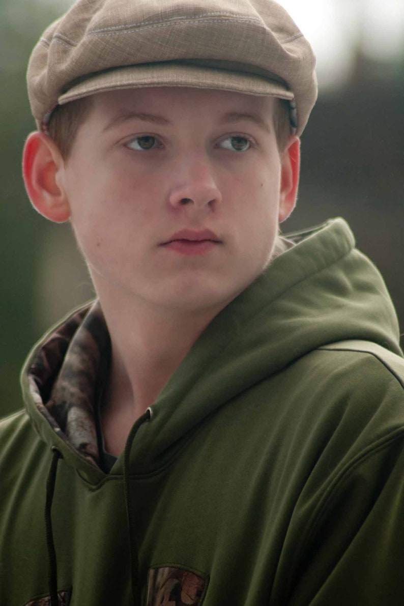 May include: A young person wearing a green hooded sweatshirt with a camouflage pattern on the inside of the hood. They are also wearing a brown newsboy cap.