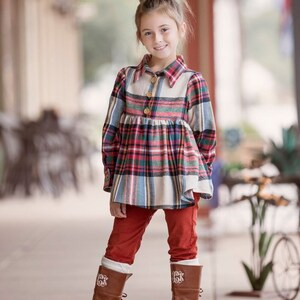 May include: A young girl wearing a red and blue plaid shirt with a white collar and red pants. She is wearing brown boots with white embroidery on the top. The girl is standing on a sidewalk in front of a building.