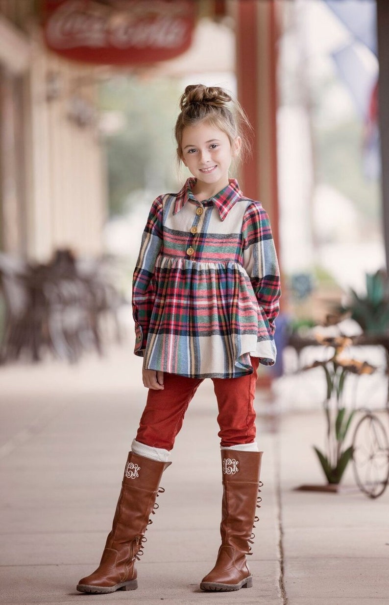 May include: A young girl wearing a red and blue plaid shirt with a white collar and red pants. She is wearing brown boots with white embroidery on the top. The girl is standing on a sidewalk in front of a building.