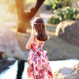 May include: A young girl wearing a floral print dress and a flower crown stands by a pond. The sun is shining brightly behind her, creating a warm glow.