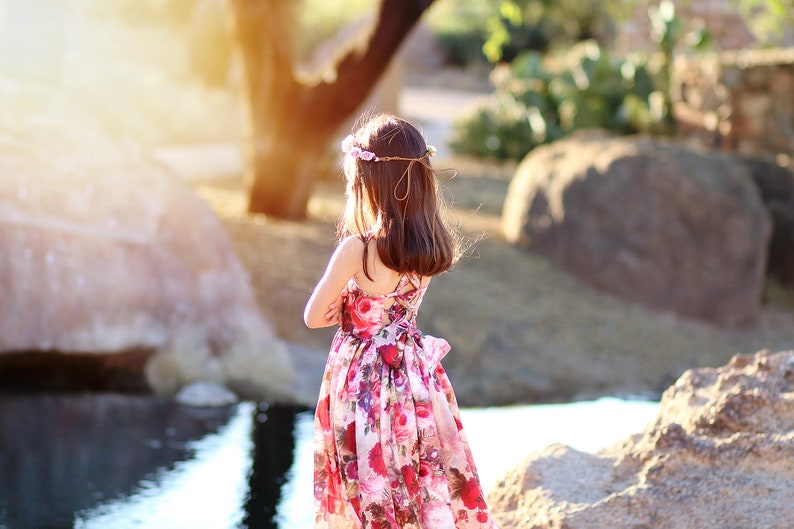 May include: A young girl wearing a floral print dress and a flower crown stands by a pond. The sun is shining brightly behind her, creating a warm glow.