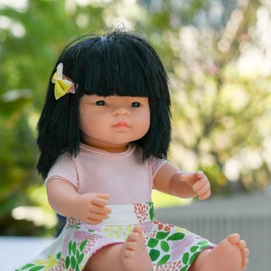 May include: A doll with black hair and a pink shirt is sitting on a white surface. The doll is wearing a colorful skirt with green, pink, and yellow flowers. The doll has a yellow bow in her hair.