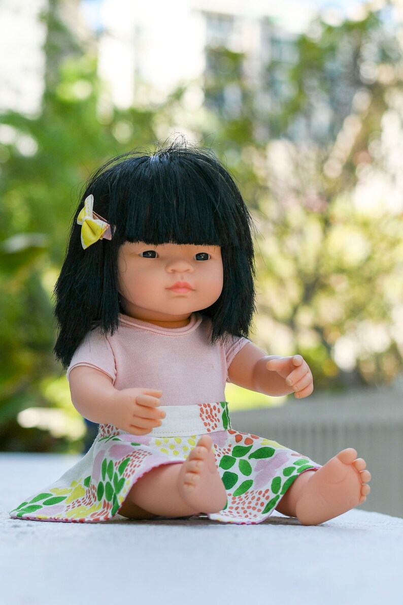 May include: A doll with black hair and a pink shirt is sitting on a white surface. The doll is wearing a colorful skirt with green, pink, and yellow flowers. The doll has a yellow bow in her hair.