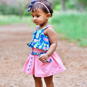 Può includere: Una bambina che indossa un vestito rosa e blu con stampa di ananas e anguria. Indossa un fiocco blu nei capelli e sandali bianchi.