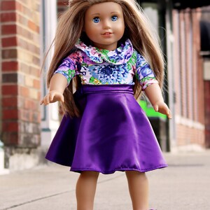 May include: A doll with long brown hair wearing a purple skirt and a floral print top with a white collar.