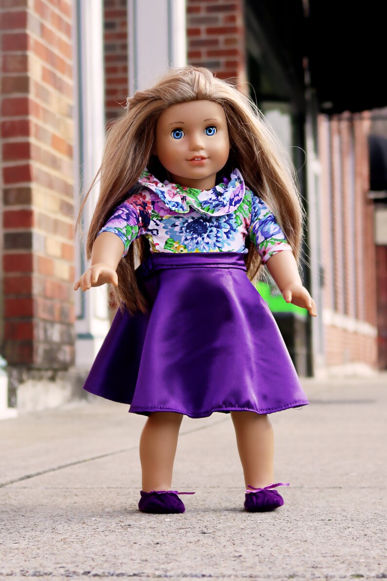 May include: A doll with long brown hair wearing a purple skirt and a floral print top with a white collar.