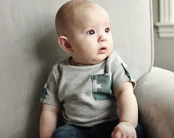 May include: A baby boy wearing a gray short-sleeved shirt with a green star pocket and blue jeans sits on a gray armchair.