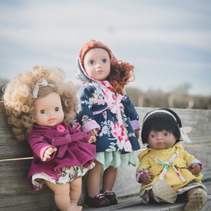 May include: Three dolls wearing different patterned coats sit on a wooden bench. The doll on the left is wearing a pink coat, the doll in the middle is wearing a blue and floral coat, and the doll on the right is wearing a yellow coat.