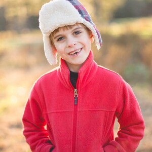 May include: A young boy wearing a red fleece jacket with a zipper and a white and red plaid winter hat. The boy is smiling and looking at the camera.