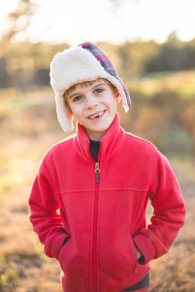 May include: A young boy wearing a red fleece jacket with a zipper and a white and red plaid winter hat. The boy is smiling and looking at the camera.