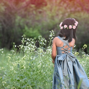 May include: A young girl wearing a light blue dress with a bow in the back and a flower crown stands in a field of white flowers.