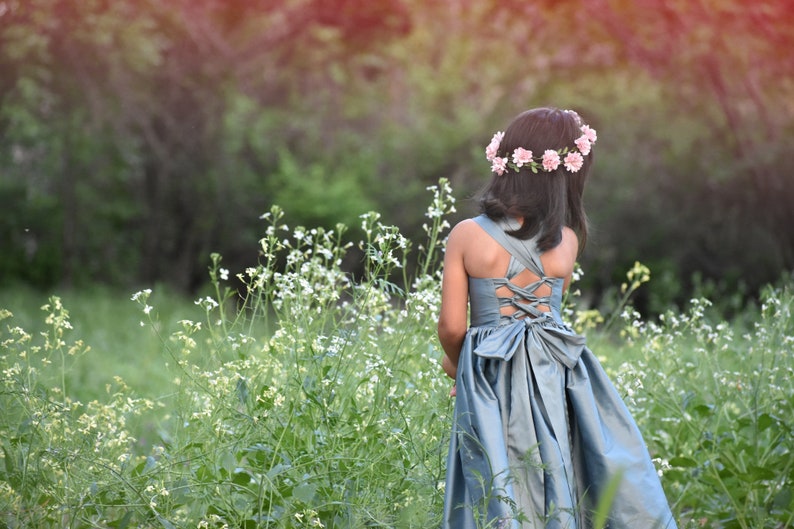 May include: A young girl wearing a light blue dress with a bow in the back and a flower crown stands in a field of white flowers.