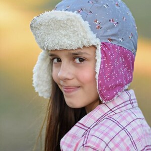 May include: A young person wearing a gray and pink patterned winter hat with a white faux fur brim and ear flaps. The hat has a small brim and is worn over the person's long brown hair. The person is wearing a pink and white plaid shirt.