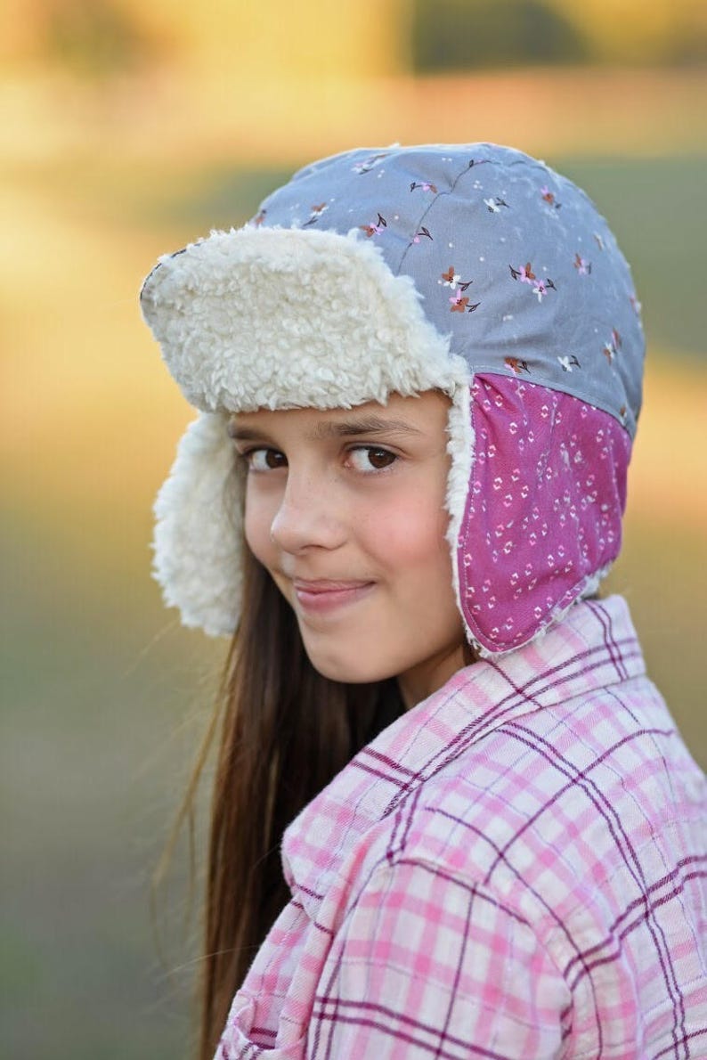 May include: A young person wearing a gray and pink patterned winter hat with a white faux fur brim and ear flaps. The hat has a small brim and is worn over the person's long brown hair. The person is wearing a pink and white plaid shirt.
