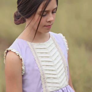 Puede incluir: Una niña pequeña con un vestido lavanda con ribete de encaje blanco y un corpiño blanco plisado. El vestido tiene un escote con volantes y una falda fruncida.