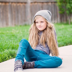 May include: A young person wearing a gray beanie, a gray long-sleeved shirt, and blue jeans sits on a concrete surface. They are wearing dark brown shoes with velcro straps.