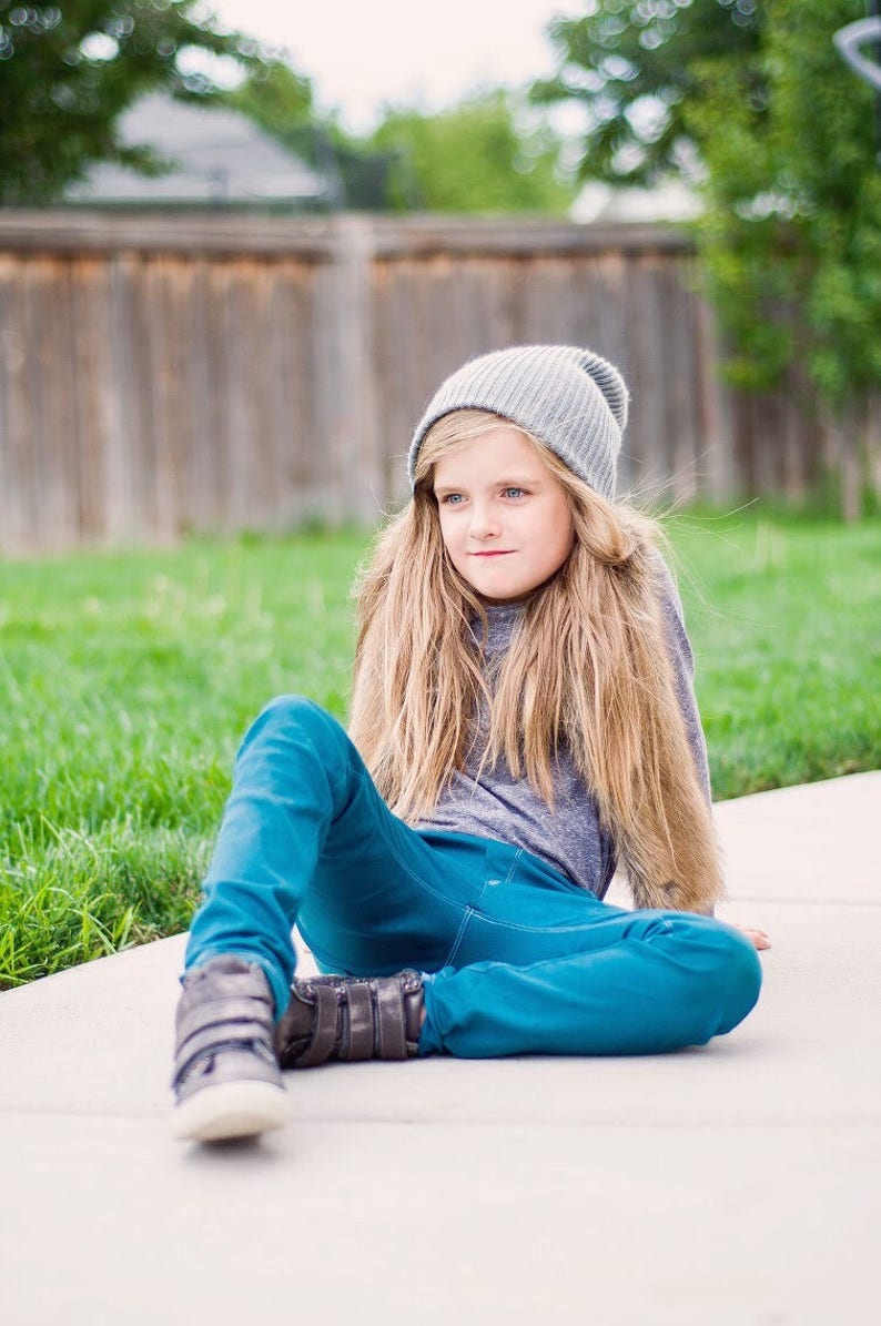 May include: A young person wearing a gray beanie, a gray long-sleeved shirt, and blue jeans sits on a concrete surface. They are wearing dark brown shoes with velcro straps.