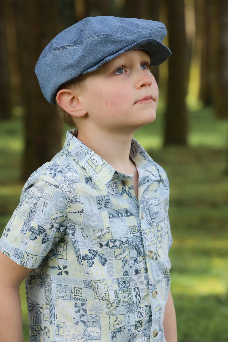 May include: A young person wearing a light blue patterned shirt and a denim newsboy cap. The person is looking up and to the right.