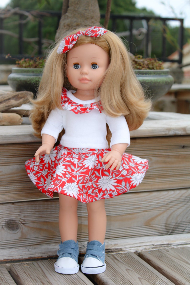 May include: A doll with long blonde hair wearing a white shirt with a red and white ruffled collar, a red and white floral skirt, and blue denim sneakers.