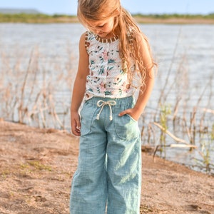 May include: A young girl wearing a white floral tank top and light blue linen pants. She is standing on a sandy beach with a body of water in the background.