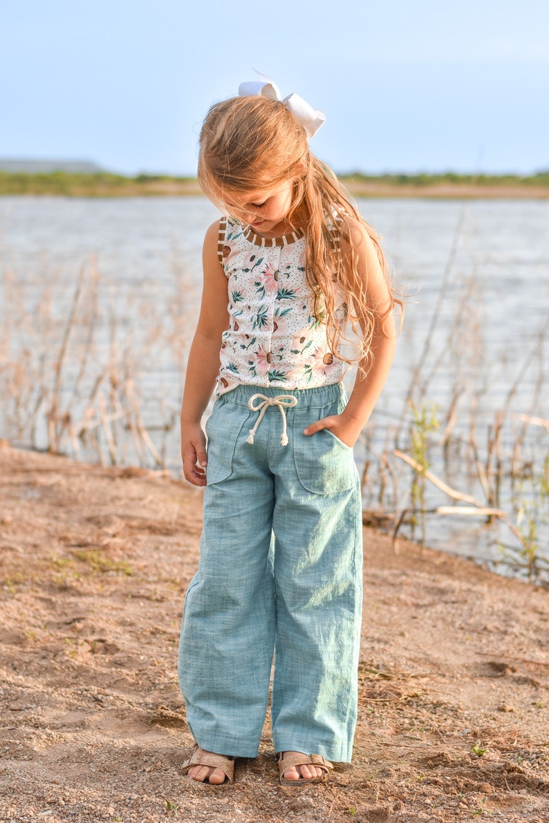 May include: A young girl wearing a white floral tank top and light blue linen pants. She is standing on a sandy beach with a body of water in the background.