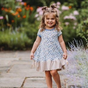 May include: A young child wearing a blue floral print dress with a light blue trim and a beige ruffle hem. The child is also wearing white shoes and has her hair styled in two buns. The background features a garden with flowers and greenery.