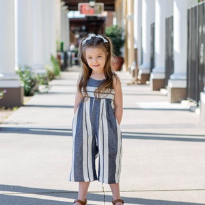 May include: A young girl wearing a blue and white striped jumpsuit with brown sandals. She is standing in a walkway with white columns on either side.