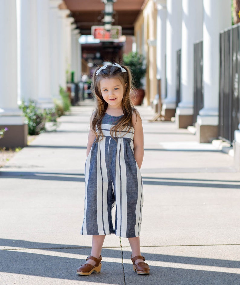 May include: A young girl wearing a blue and white striped jumpsuit with brown sandals. She is standing in a walkway with white columns on either side.