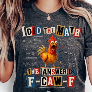 May include: A dark heather gray short-sleeve t-shirt featuring a graphic of a surprised yellow chicken surrounded by mathematical equations. The text reads I DID THE MATH and THE ANSWER IS F-CAW-F in a colorful ransom note style font.
