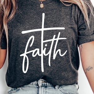 May include: Dark gray t-shirt with the word "faith" in white script and a cross design. The shirt is a crew neck style and appears to be made of a soft material. The text is centered on the front of the shirt.