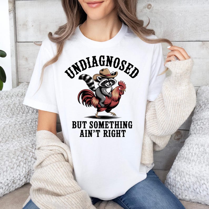 May include: White t-shirt featuring a graphic of a raccoon wearing a cowboy hat riding a rooster. The text on the shirt reads "UNDIAGNOSED BUT SOMETHING AIN'T RIGHT" in a bold, black font. The design is centered on the front of the shirt.