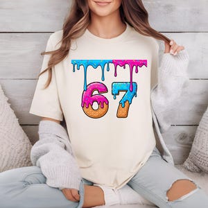 May include: Cream-colored t-shirt featuring the number "67" in a colorful, dripping ice cream design. The numbers are pink and blue, with a waffle cone texture at the base. The top of the design has blue and pink drips.