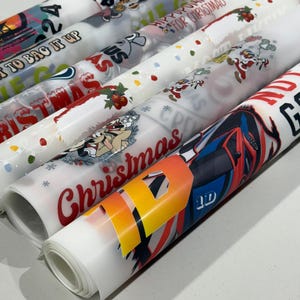 May include: Rolled sheets of printed wrapping paper with various designs, including Christmas themes, cartoon characters, and abstract patterns. The colors include red, white, blue, and yellow. Some sheets have text like "Christmas" and "To Bag It Up".