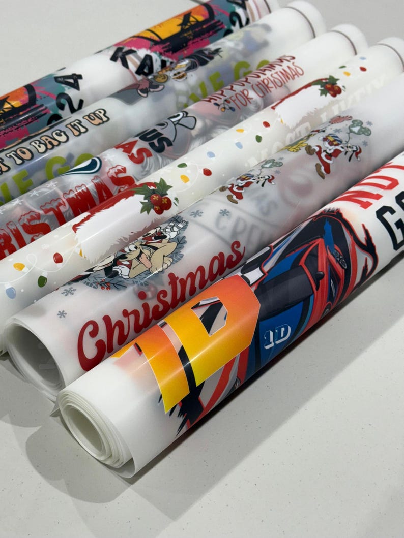 May include: Rolled sheets of printed wrapping paper with various designs, including Christmas themes, cartoon characters, and abstract patterns. The colors include red, white, blue, and yellow. Some sheets have text like "Christmas" and "To Bag It Up".