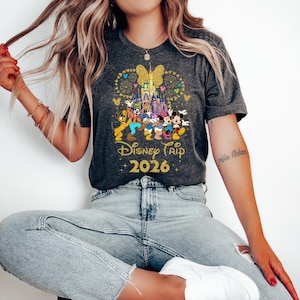 May include: Dark gray t-shirt featuring a colorful graphic of Disney characters in front of a castle, with fireworks and a butterfly design. The text "Disney Trip 2026" is in gold. The shirt is short-sleeved.